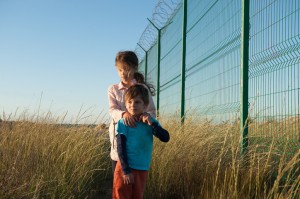 two caring family poor kids boy and girl in dirty clothes near fencing with barbed wire among high grass on state border